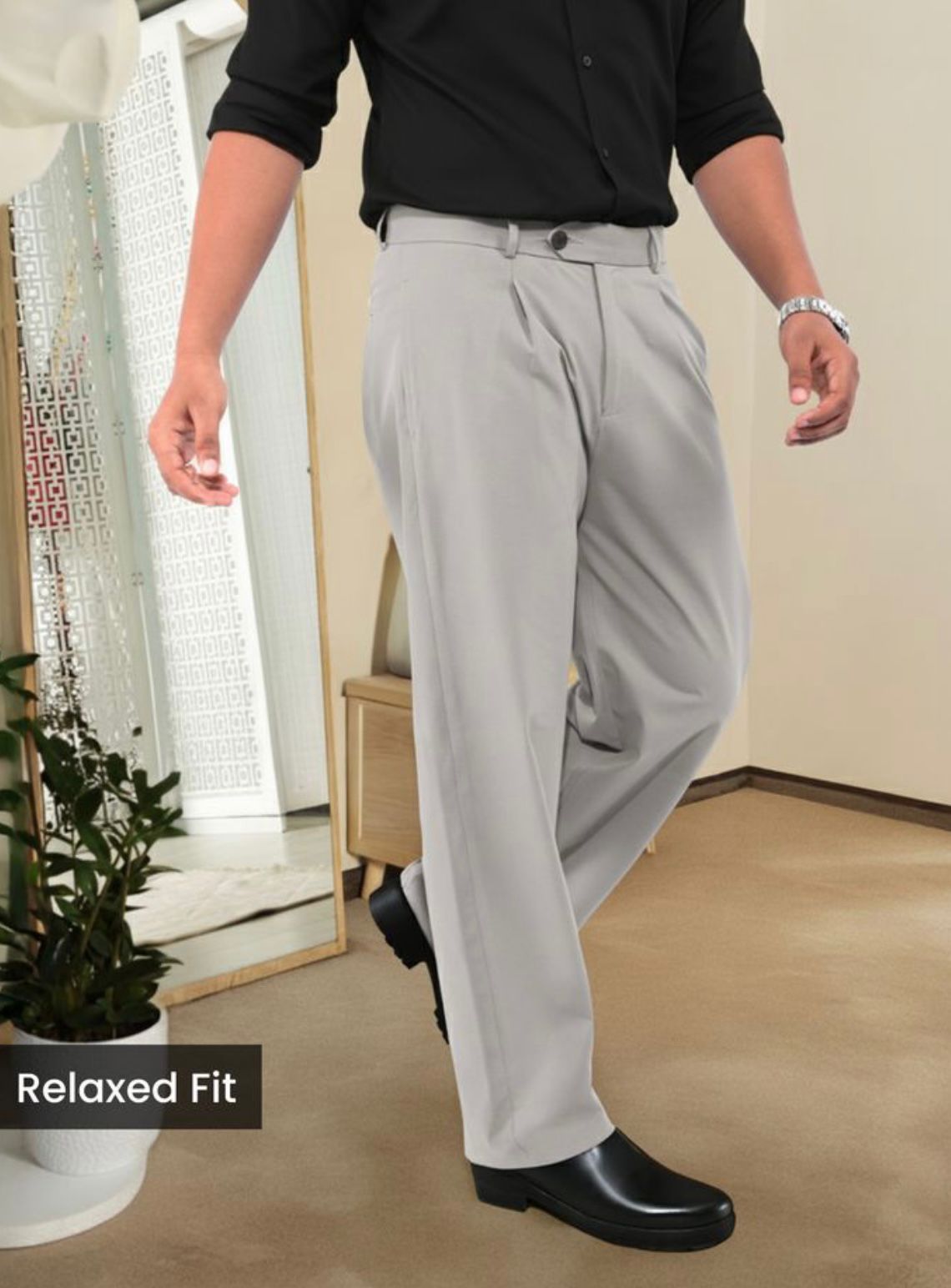 Man wearing light grey relaxed fit pleated trousers with a black shirt and dress shoes, standing indoors